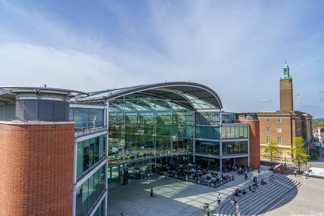 The Forum in Norwich: a curved glass atrium between brick wings, with people sitting on the steps of Millennium Plain and City Hall's tower behind.